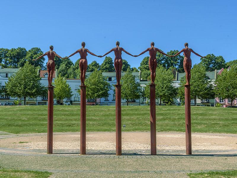 Portishead Economic Development Fund: Sculpture of five human figures holding hands while balancing on tall, narrow pillars, set in a grassy area with trees and buildings in the background.
